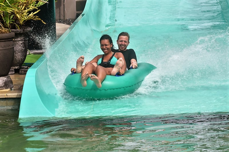 2 people coming down a green slide on a green inflatable. Both of them are very happy. Is travelling a hobby? i reckon so.